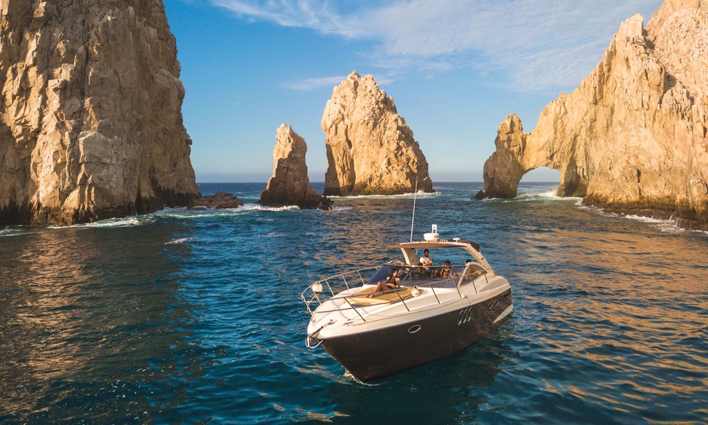 a boat in the water with rocks in the background with Arch of Cabo San Lucas in the background